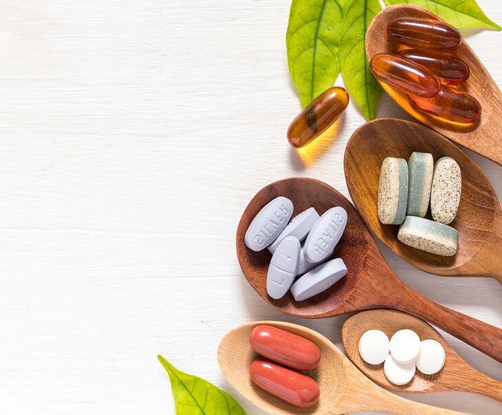Variety of vitamin pills in wooden spoon on white background with green leaf, supplemental and healthcare product, flat lay surface